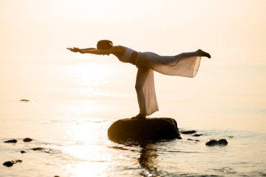 Side view of woman silhouette doing yogas balancing stick pose standing on stone at beach at morning. Tuladandasana.