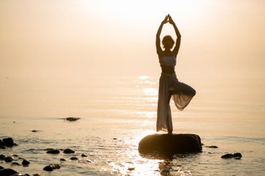 Silhouette of woman who practices yogas tree pose on the beach standing on stone at sunset. Healthy lifestyle concept.
