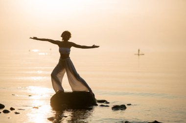 Silhouette of beautiful woman practicing yoga or pilates at sunset standing in Warrior pose on stone.