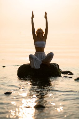 pretty young woman with closed eyes practicing yoga meditate in lotus pose outdoors sitting on stone at water. Healthy life concept.