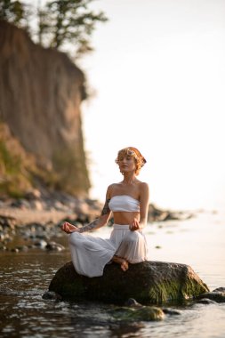 Attractive woman with closed eyes practicing yoga meditate in lotus pose outdoors sitting on stone at water. Healthy life concept.