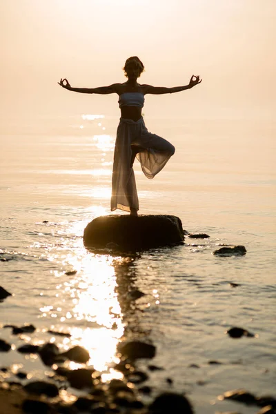great view of woman silhouette who practices yogas tree pose on the beach standing on stone at sunset. Healthy lifestyle concept.