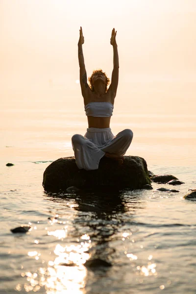 pretty young woman with closed eyes practicing yoga meditate in lotus pose outdoors sitting on stone at water. Healthy life concept.