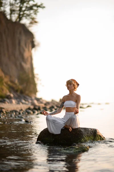 woman with closed eyes practicing yoga meditate in lotus pose outdoors sitting on stone at water. Healthy life concept.