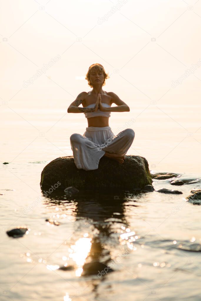 pretty woman with closed eyes practicing yoga meditate in lotus pose outdoors sitting on stone at water. Healthy life concept.