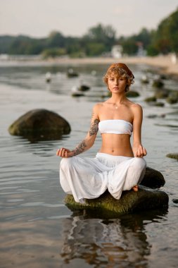 Woman practices yoga position doing meditation in tranquil outdoor sitting on stone in water. Mental wellbeing healthy lifestyle.