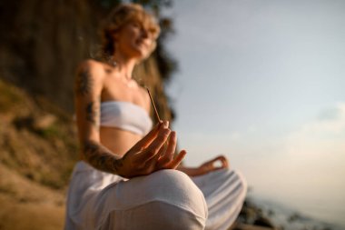 selective focus on steaming incense stick in the hand of young woman meditating in yoga pose on the beach