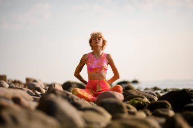 Woman practices yoga position doing meditation in tranquil outdoor sitting on stones at coast. Mental wellbeing healthy lifestyle.