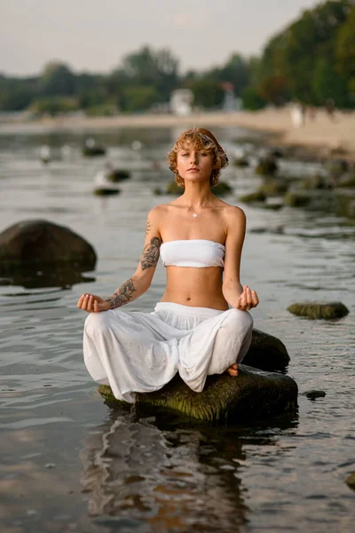 calm peaceful attractive healthy young woman doing yoga meditation sitting on big stone in water. Relaxation yoga exercise concept.