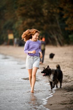 Barefoot young woman having fun and runs with the fur dog along the shore. Outdoor leisure activity