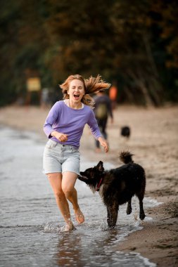 Fur dog biting running barefoot young woman along the shore. Games of owners and dogs in nature