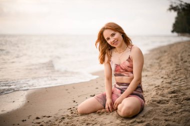 Great view on caucasian smiling beautiful red-haired young woman sitting on sand of beach. Blurred background