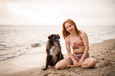 cute dog and smiling young pretty woman are sitting on the sandy shore of the beach. Pets and owners
