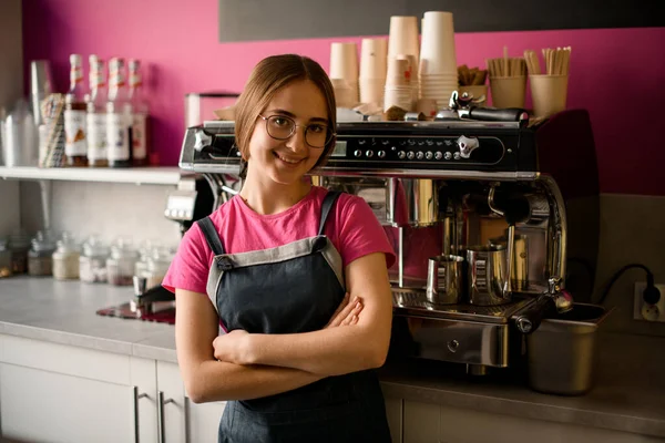 beautiful young female barista in glasses stands with her arms crossed on her chest in coffee shop and professional coffee machine in the background