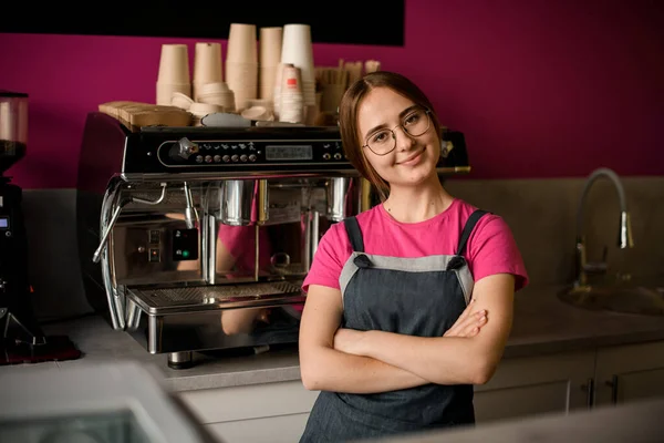 portrait of beautiful young woman barista in glasses with her arms crossed on her chest in coffee shop. Professional coffee machine in the background