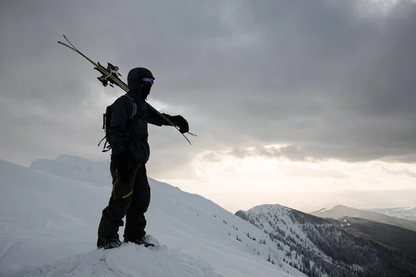 Narciarz Kombinezonie Narciarskim Nartami Ramionach Stoi Pokrytym Śniegiem Szczycie Góry — Zdjęcie stockowe