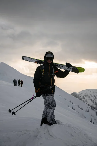 male tourist in black ski suit and with backpack with skies on his shoulders on snow-covered mountain. Ski touring and freeride concept