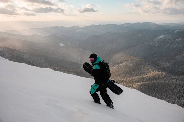 スノーボードを手にスキースーツを着た男が美しい山の風景を背景に雪に覆われた斜面を歩く — ストック写真