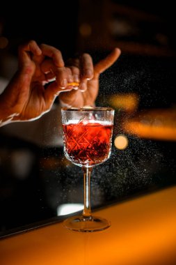 Barmenin narenciye kabuğundan su sıçrattığı alkollü bir içecekle kristal bardağa odaklan.