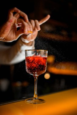 Close-up of glass with alcoholic cocktail on bar while hands of male bartender sprinkle by citrus zest juice on it