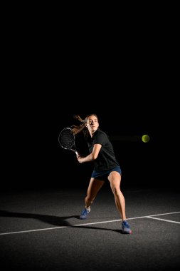 Genç bayan tenis oyuncusuna raketle vurmaya hazır sarı tenis topuna vurmaya hazır. Koyu arkaplan.