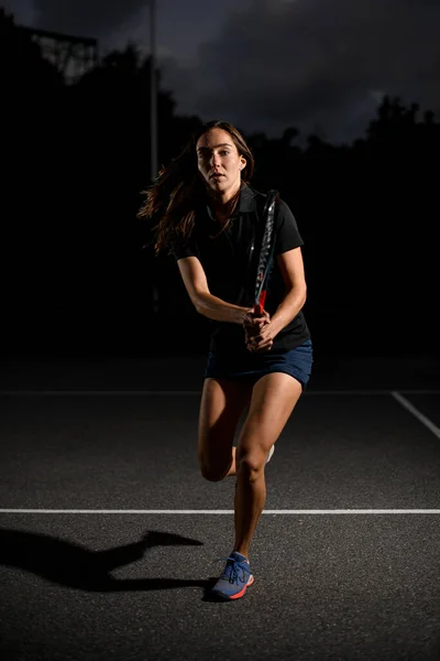 屋外テニスコートでテニスラケットで走る美しい現役女子テニス選手の姿 暗い背景 — ストック写真