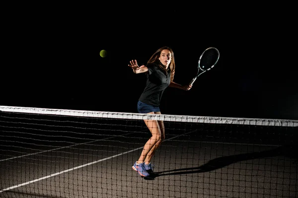 空中で黄色のテニスボールの素晴らしいショットとそれを打つ準備ができて彼女の手にラケットを持つ女性のテニス選手 暗い背景 — ストック写真