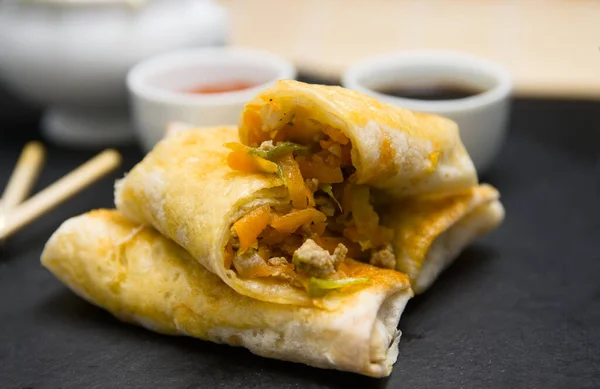 Close-up of some homemade spring rolls on a black slate plate with soy sauce, sweet and sour and some Chinese wooden chopsticks