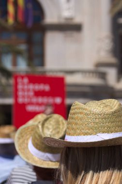 Seyirciler arasında tanınmayan bir kadının, herkesin güneşten korunmak için hasır şapka taktığı bir açık hava etkinliğinde yakın plan fotoğrafı.