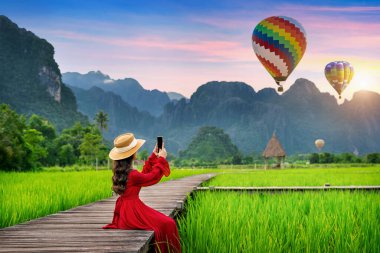 Ahşap yolu üzerinde oturan genç kadın ve Vang Vieng, Laos yeşil pirinç alanıyla balonlar bak.