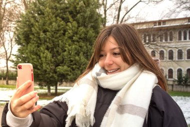 Genç bir kadın akıllı telefonuyla selfie çekiyor. Genç bir kadın cep telefonuyla video görüşmesi yapıyor. Sıcak elbiseli bir kadın halka açık bir parkta gülümsüyor ve telefonuyla oynuyor..