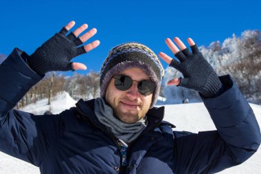 Güneş gözlüklü, kalın elbiseli genç adam kış mevsiminde dağın tadını çıkarıyor. Halka açık parkta kollarını açıp gülümseyen neşeli bir adam..