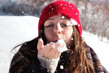 Kırmızı bereli neşeli kız dağda ellerinden kar üflüyor. Kırmızı şapkalı, karı uçuran kıza odaklan. Elinde kar tutan neşeli genç kız. Karda eğleniyoruz.