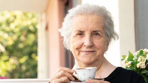 Old smiling woman looking at camera, drinking turkish coffee and relaxing on balcony. Carefree senior woman in 70s smiling and enjoying the time.