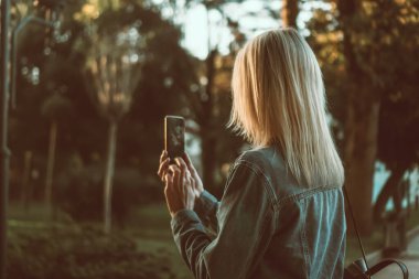 Kot ceketli genç sarışın kadın parktaki güzel bir manzaranın cep telefonuyla fotoğraf çekiyor, arkadan. Mobil fotoğrafçılık, seyahat kavramı