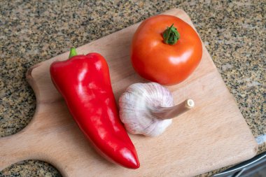 Ahşap bir mutfak tabağında domates, kırmızı biber ve bir baş sarımsak var.