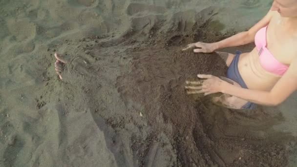 한 여자가 검은 모래와 해변에 앉아서 자신을 묻으려고 하고 있습니다. — 비디오