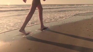İnce seksi kadın bacakları kumlu bir sahilde deniz suyunun dalgalarında yürüyor..