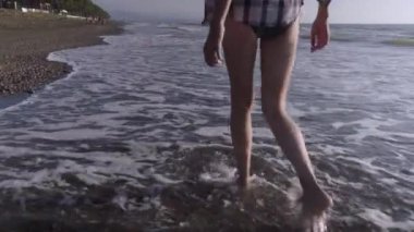 İnce seksi kadın bacakları kumlu bir sahilde deniz suyunun dalgalarında yürüyor..
