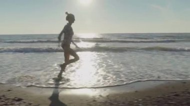 İnce bir kadın koşar ve kumlu bir sahilde deniz suyunun dalgalarına atlar..