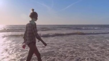 İnce bir kadın kumlu bir sahilde deniz suyunun dalgalarında yürüyor..