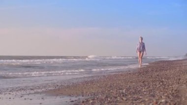 İnce bir kadın kumlu bir sahilde deniz suyunun dalgalarında yürüyor..