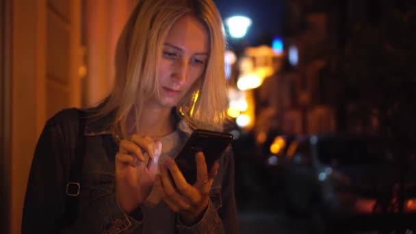 Μια ξανθιά με τζιν μπουφάν τριγυρνάει στην πόλη κοιτάζοντας το κινητό της. — Αρχείο Βίντεο