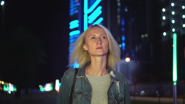 Egy fiatal, gyönyörű, karcsú szőke a kamerába megy az éjszakai városban. — Stock videók