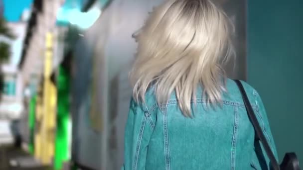 Vista trasera de la joven mujer con estilo en chaqueta de mezclilla caminando por el centro de la ciudad — Vídeos de Stock