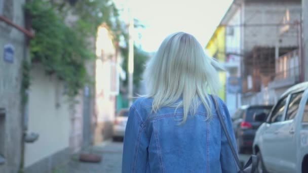 Zadní pohled na mladé stylové blondýny v džínové bundě procházky centrem města — Stock video