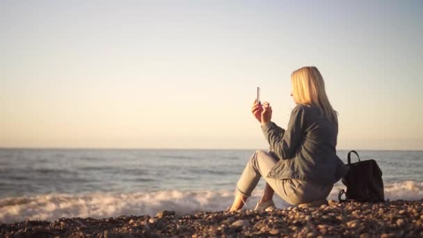 Una mujer se sienta en la orilla del mar y escribe un atardecer en su teléfono. — Vídeos de Stock