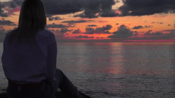 Una mujer se sienta sobre piedras junto al mar y mira una hermosa puesta de sol, — Vídeos de Stock