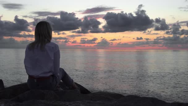 Una mujer se sienta sobre piedras junto al mar y mira una hermosa puesta de sol, — Vídeos de Stock