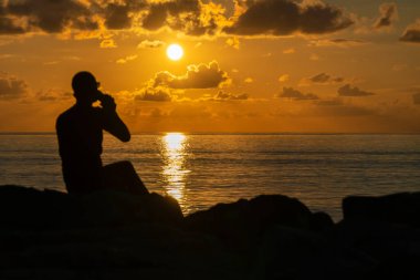 Gün batımında deniz kıyısında kahve içen bardaklı bir adamın silueti. Denizde güzel, sihirli bir gün batımı ve silueti olan bir adam. Arkadan fotoğraf, güneşe odaklan.